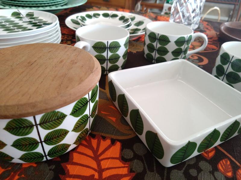 Set med porslin med bladmönster, inklusive en behållare med trälock, koppar och ett fyrkantigt fat, uppsatt på en färgglad duk.