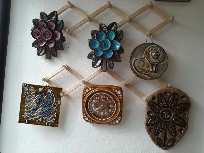En väggutställning med olika dekorativa föremål i keramik och metall, inklusive två blomformade delar, en lejonplakett, en klocka och två intrikata mönster, allt arrangerat på ett trägaller.