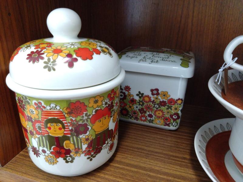 Två keramiska behållare med färgglada blommönster, en med lock och karaktärsdesign och den andra rektangulär med text. De står uppställda på en trähylla bredvid en kopp och ett fat.