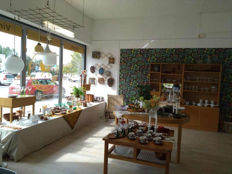 En väl upplyst butiksinredning med träbord som visar olika keramik, glas och dekorativa föremål. Hyllor står mot väggarna och stora fönster släpper in naturligt ljus. En färgglad väggtapet ger stil.
