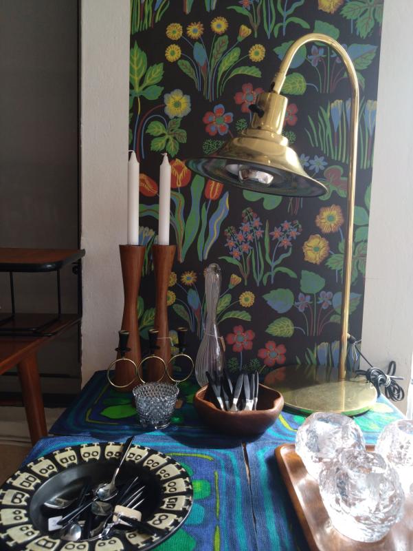 En bordsuppsättning med ljusstakar, redskap, en dekorativ skål, kristallliknande dekor och en skrivbordslampa i mässing, ställd mot en blommönstrad vägg.