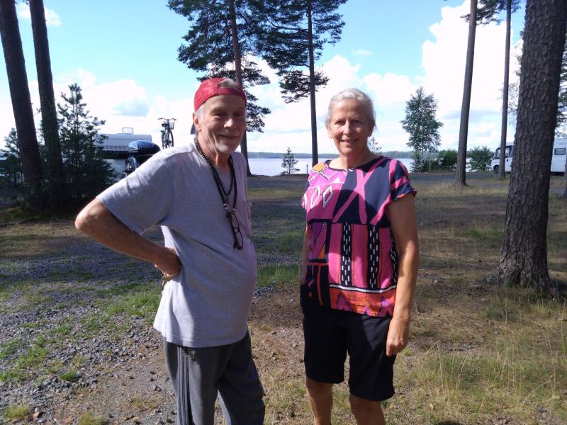 En äldre man och kvinna står utomhus på en grus- och gräsyta med träd, en sjö och husbilar i bakgrunden en solig dag.