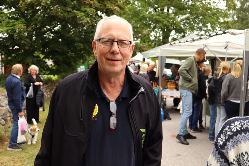 En äldre man med glasögon och svart jacka står utomhus på en marknad, med människor och stånd i bakgrunden.