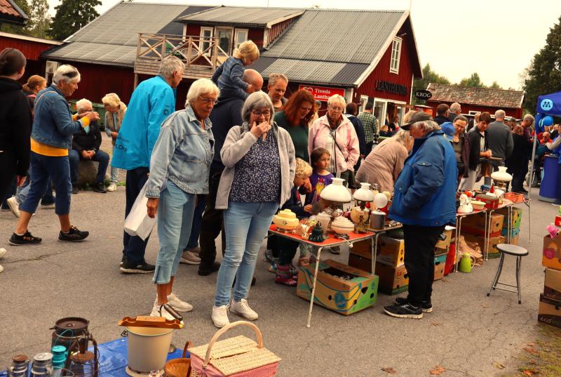 Människor tittar på olika hushållsartiklar och lampor som visas på bord på en utomhusmarknad framför träbyggnader.