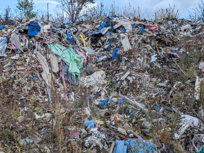 En hög med blandat skräp och avfall, bland annat plastpåsar, kläder och diverse avfallsmaterial, utspritt utomhus bland ogräs.
