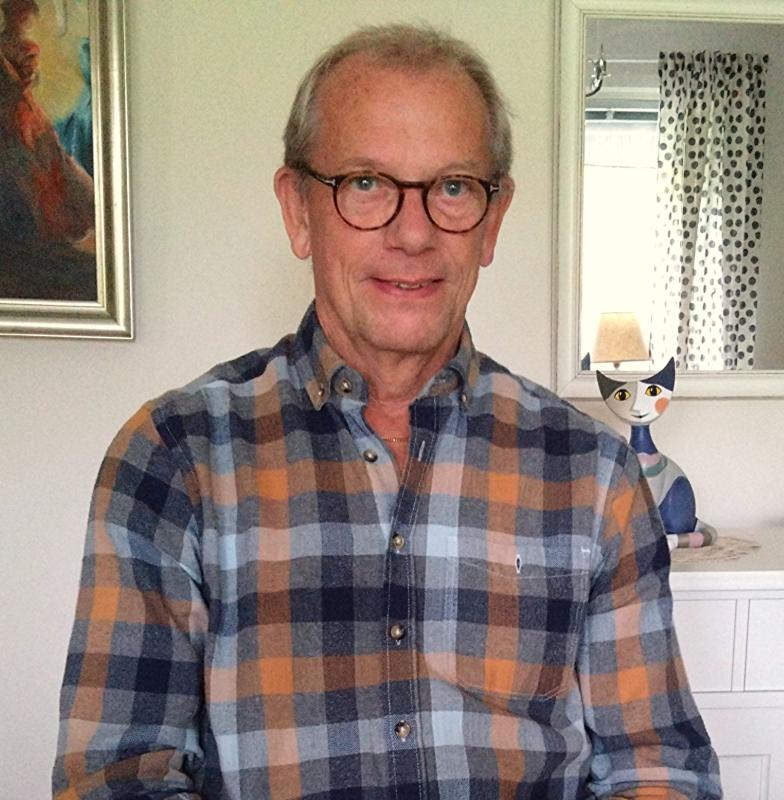 En äldre man med glasögon och rutig skjorta sitter inomhus. Bakom honom finns ett målat porträtt, en spegel, ett fönster med en prickig gardin och en dekorativ kattfigur.