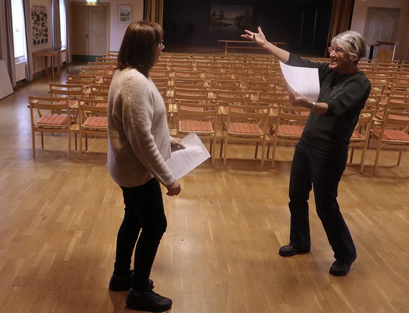 Två personer står i ett tomt rum med rader av stolar; den ena håller i papper och gestikulerar energiskt mot den andra.