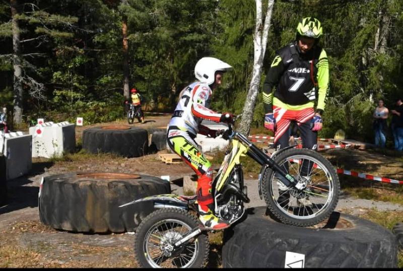 Ett barn i skyddsutrustning cyklar på en smutscykel över stora däck, guidad av en vuxen i liknande klädsel. Andra cyklister och åskådare är i bakgrunden mitt i ett skogsområde.