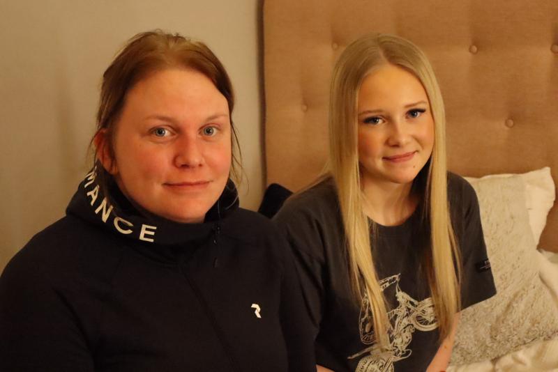 Två personer sitter på en säng, en klädd i en svart jacka och den andra en mörk t-shirt. De är inomhus, med en tuftad sänggavel som syns i bakgrunden.