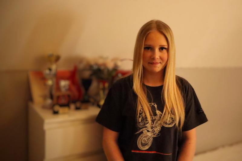 En ung kvinna med långt blont hår står inomhus framför en byrå och visar flera troféer och medaljer.