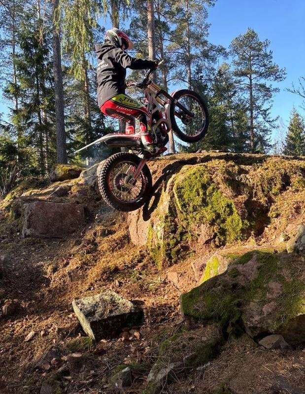 En person på en smutscykel klättrar upp i en brant, stenig terräng i ett skogsområde under klarblå himmel.