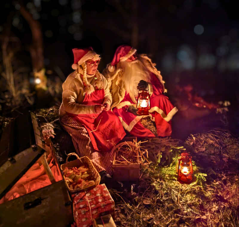 Två personer klädda i tomtedräkter sitter på marken i en skog på natten, omgivna av lyktor, en korg, inslagna presenter och en öppen kista.
