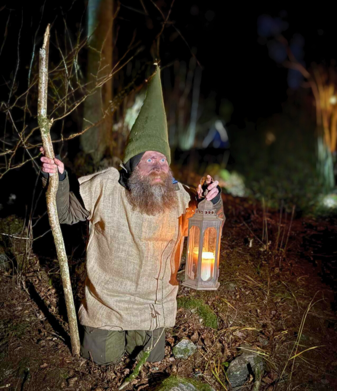 En person klädd som en tomte med en hög grön hatt, som håller i en trästav och en tänd lykta, knäböjer utomhus på natten bland träd och växter.