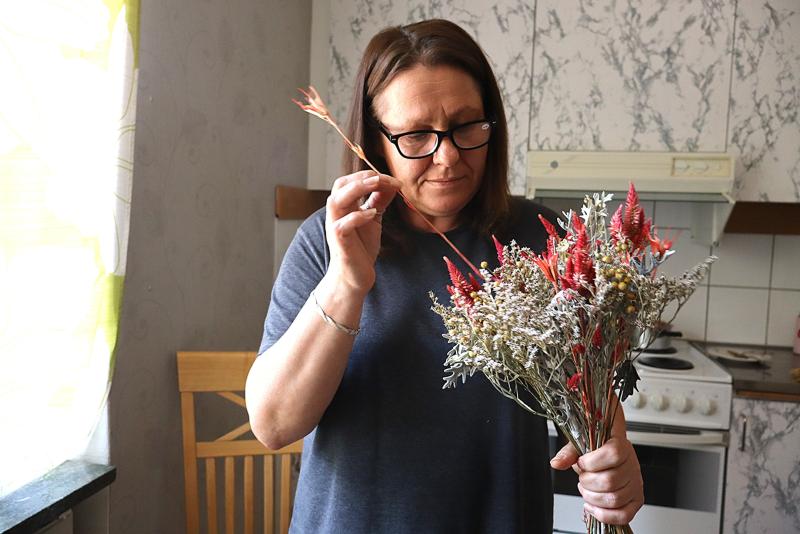 En kvinna med glasögon står i ett kök och håller en bukett torkade blommor i handen, samtidigt som hon tittar noga på en av stjälkarna.