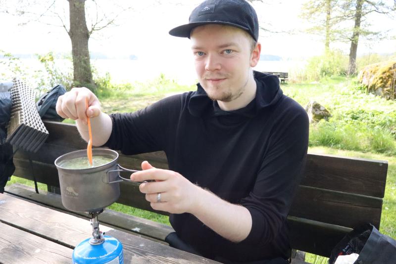 En person sitter vid ett picknickbord utomhus, lagar mat i en gryta på en bärbar spis och rör om med en gaffel.