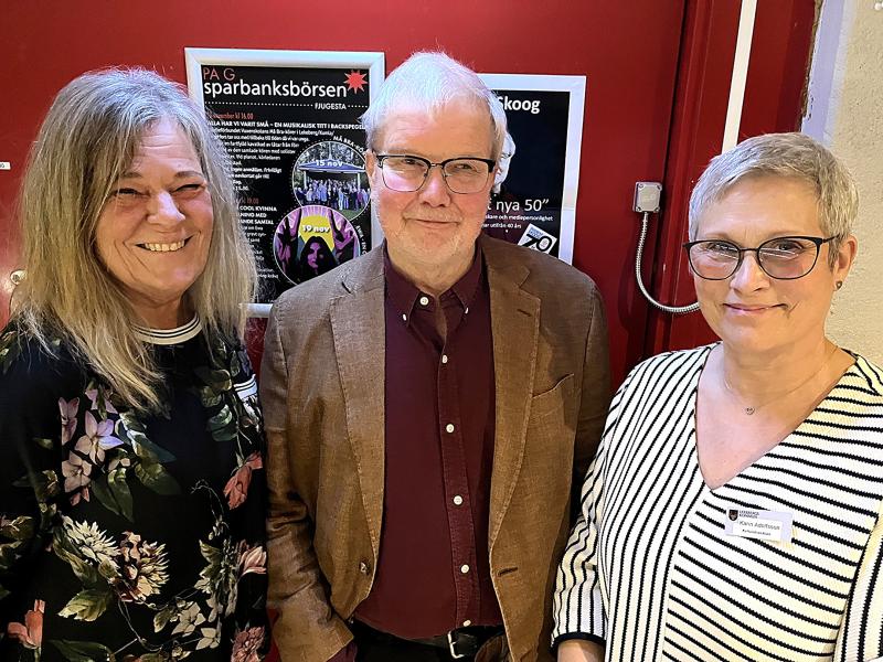 Tre vuxna står inomhus framför en röd vägg med affischer; två kvinnor står på var sin sida om en man, och alla ler mot kameran.