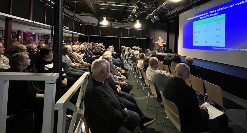 En grupp människor sitter i ett svagt upplyst auditorium och lyssnar på en person som presenterar en bild med text och en datatabell som projiceras på en stor skärm.