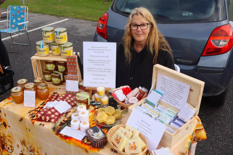 En kvinna sitter vid en marknadsbod utomhus där hon har ställt fram burkar med honung, bivaxprodukter, tvålar och informationsskyltar på en mönstrad duk framför en parkerad bil.