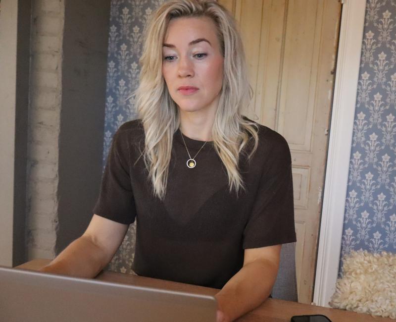 En kvinna med långt blont hår sitter vid ett bord och fokuserar på en bärbar dator framför sig. En smartphone ligger på bordet.