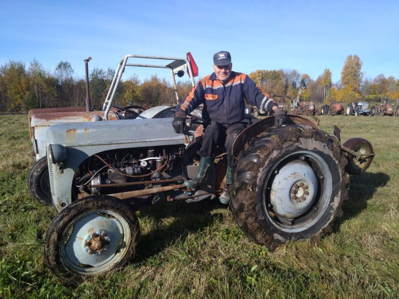 En person som sitter på en gammal traktor i ett gräsbevuxet fält med träd i bakgrunden.