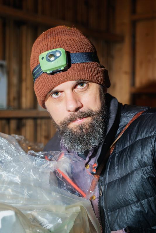 En man med en brun stickad mössa och en pannlampa står inomhus och tittar in i kameran. Han har skägg och håller i en stor plastpåse.
