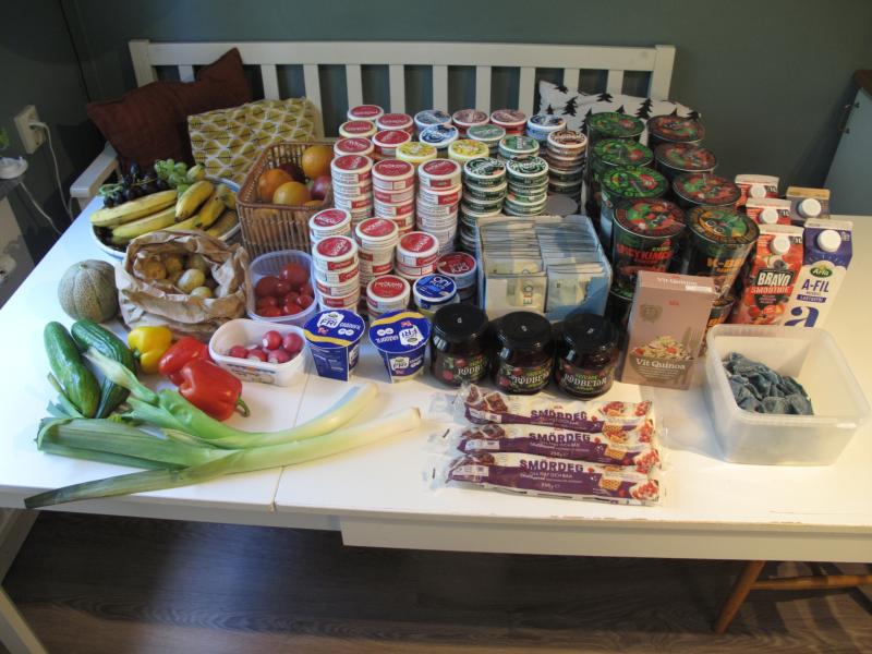 Ett köksbord fullt av olika livsmedel, bland annat yoghurtbägare, grönsaker, frukt, juiceförpackningar, förpackad ost, chokladkakor och burkar. Varorna är prydligt uppställda i rader.