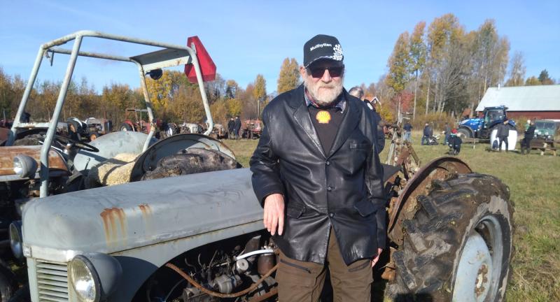 En man i en svart skinnjacka och keps står bredvid en vintagetraktor på ett fält med höstträd i bakgrunden.