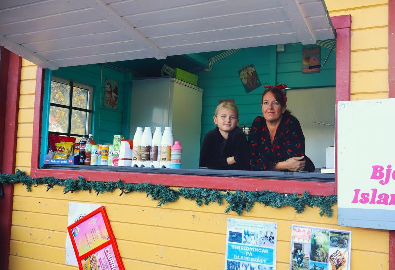 Ett barn och en vuxen står bakom disken i en färgglad kiosk där olika flaskor, snacks och affischer är utställda.