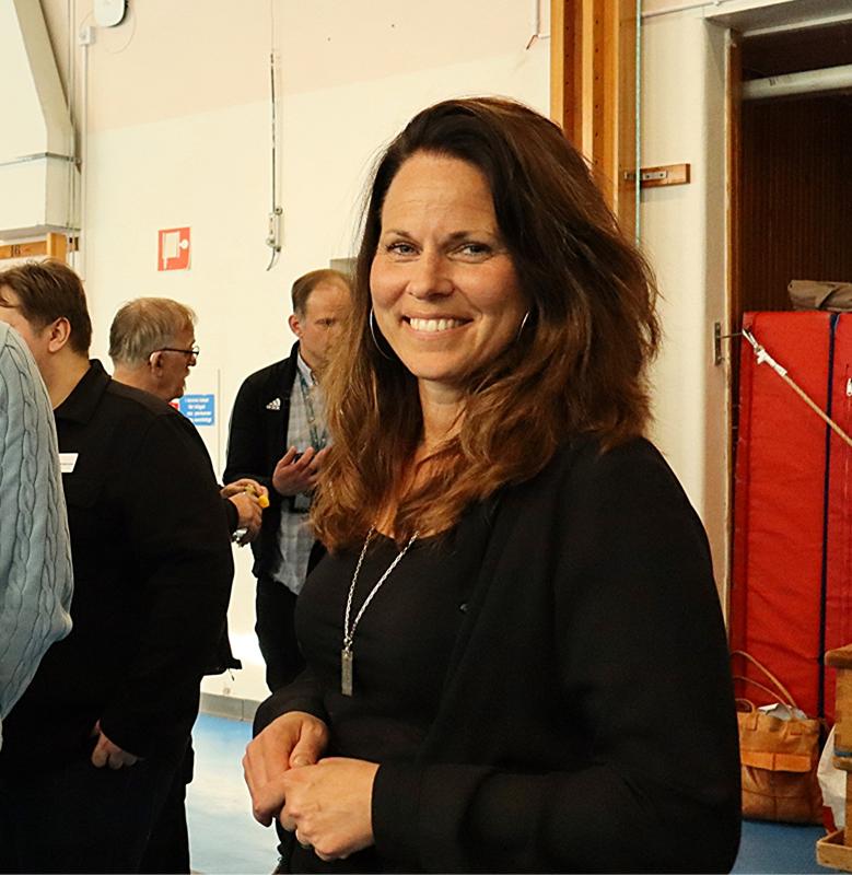 En kvinna med långt brunt hår, klädd i svart topp och halsband, står inomhus och ler mot kameran. Andra människor syns i bakgrunden.