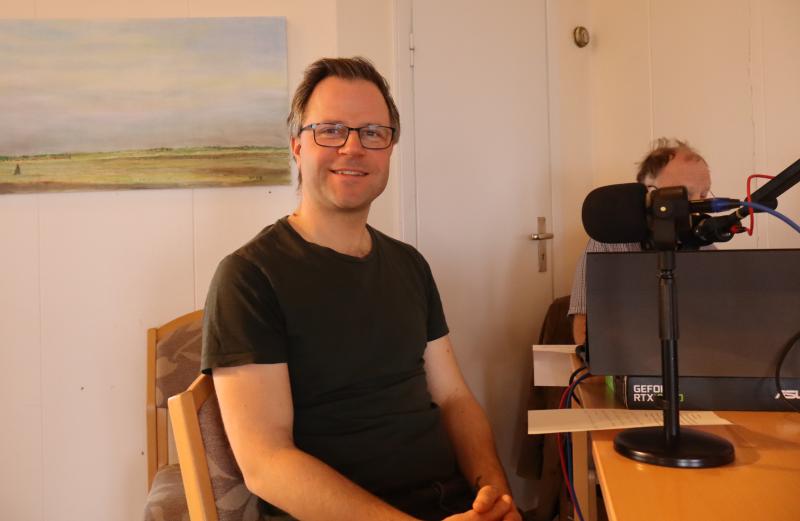 En man med glasögon och mörk t-shirt sitter nära en mikrofon och datorutrustning i ett rum, med en annan person delvis synlig i bakgrunden.