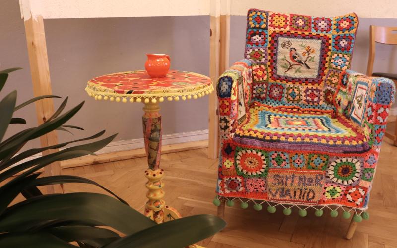 En färgglad fåtölj med virkade överdrag och ett matchande runt sidobord med en liten orange vas på, placerade på ett trägolv bredvid en växt.