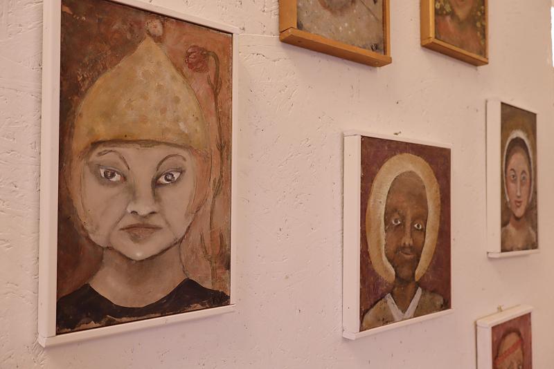 En samling inramade porträttmålningar med stiliserade, uttrycksfulla ansikten hänger på en vit vägg med textur i ett konstgalleri.