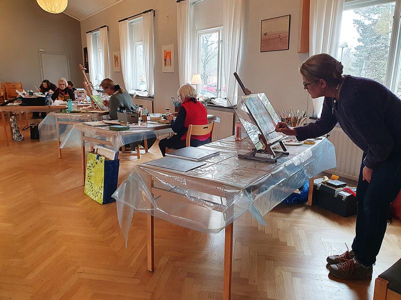 Människor sitter vid bord och målar i ett väl upplyst rum, med en person som står och arbetar vid ett staffli i förgrunden.
