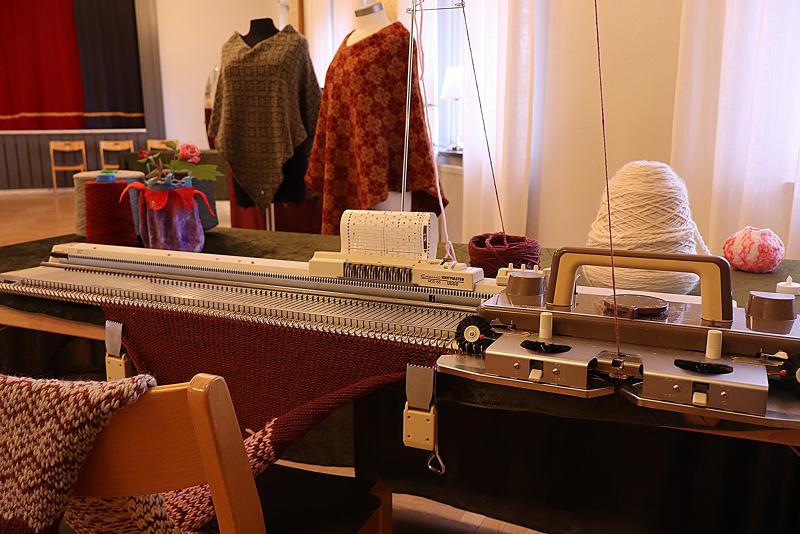 En stickmaskin med delvis färdigt tyg, garnkoner och färdiga stickade plagg utställda på skyltdockor och bord i ett väl upplyst rum.
