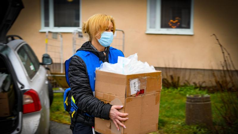 En person iförd mask och blå väst bär en stor kartong fylld med förnödenheter utomhus i närheten av en byggnad.