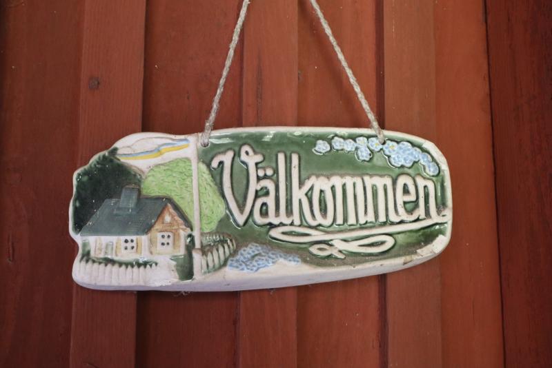 På en röd trävägg hänger en keramikskylt med ett hus och blommor som illustration och det svenska ordet 