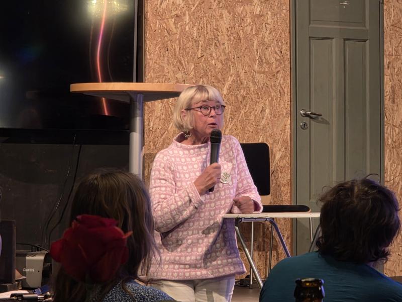 En äldre kvinna med grått hår och glasögon talar i en mikrofon medan hon sitter ner och vänder sig till en liten publik i en inomhusmiljö.