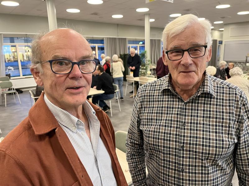 Två äldre män med glasögon står inomhus på en social tillställning, med flera personer som sitter och pratar i bakgrunden.