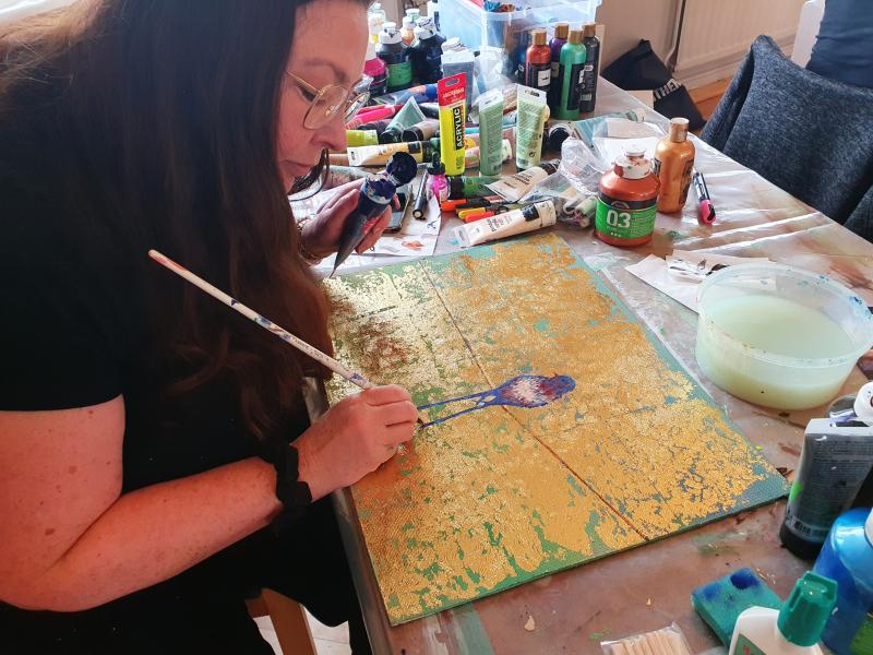 En kvinna med långt brunt hår målar på en duk med guldtextur vid ett rörigt bord fyllt med konsttillbehör, färgtuber och en skål med vatten.