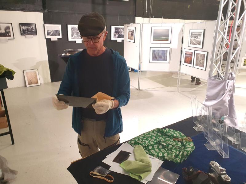 En man med handskar undersöker ett fotografi eller en plåt i ett konstgalleri, omgiven av inramade foton och fotoutrustning på ett bord.