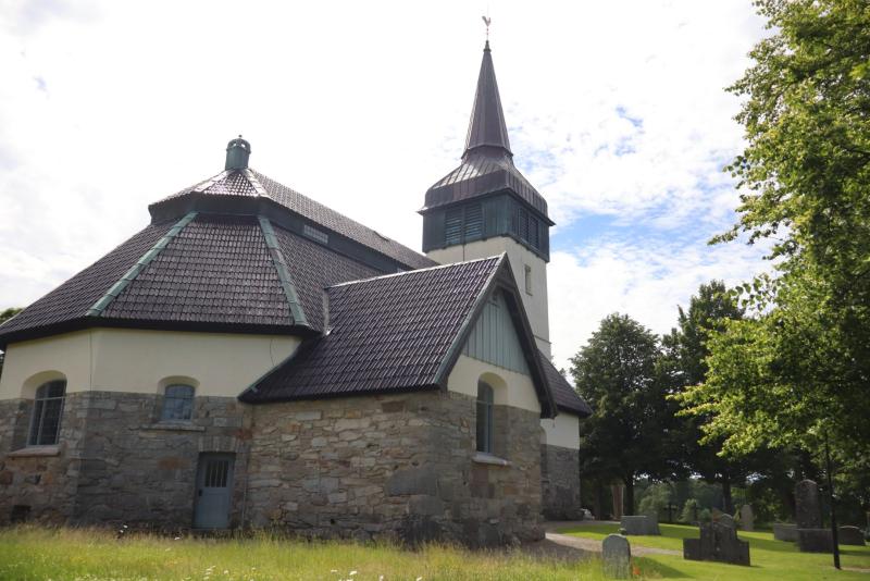 En stenkyrka med ett högt torn, omgiven av gräs och gravstenar, med träd i bakgrunden och en delvis molnig himmel ovanför.