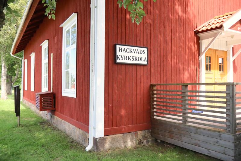 En röd träbyggnad med vita lister, med en skylt vid ingången där det står ”Hackvads Kyrkskola”, med gräs och träd i närheten.