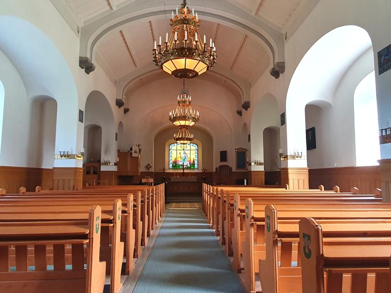 Interiören i en kyrka med träbänkar, ljuskronor, välvt tak och ett målat glasfönster ovanför altaret längst bak.