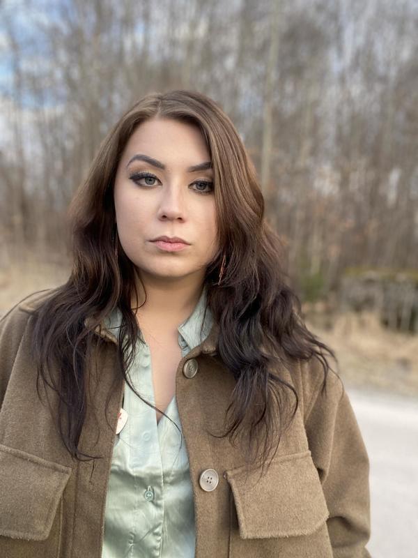 En person med långt mörkt hår iklädd brun jacka och ljusblå skjorta, stående utomhus med kala träd i bakgrunden.