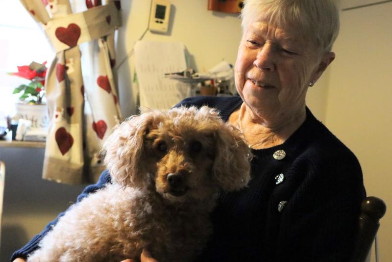 En äldre kvinna som sitter inomhus och håller en liten lockig hund i knät. Bakom dem syns delvis en fönstergardin med hjärtmönster.