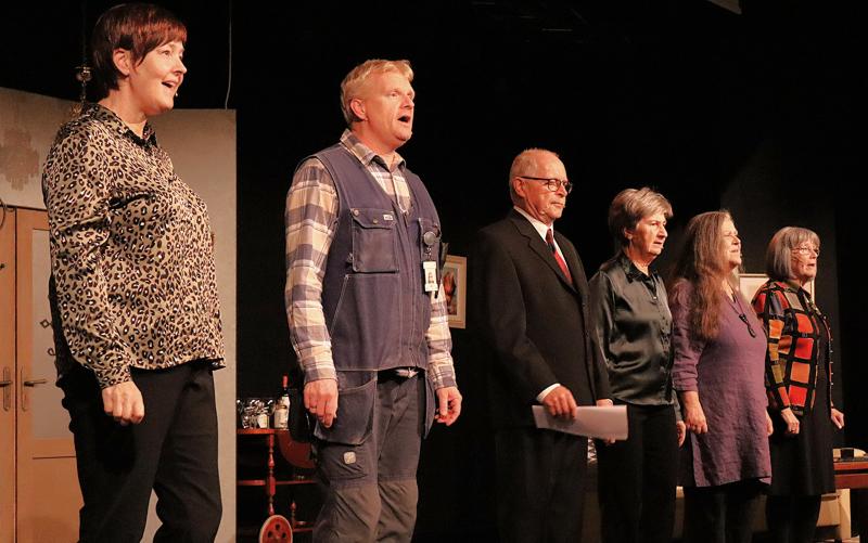 Fem vuxna står på rad på scenen, vända framåt, klädda i olika utstyrslar, med scenrekvisita och en målad fond synlig bakom sig.