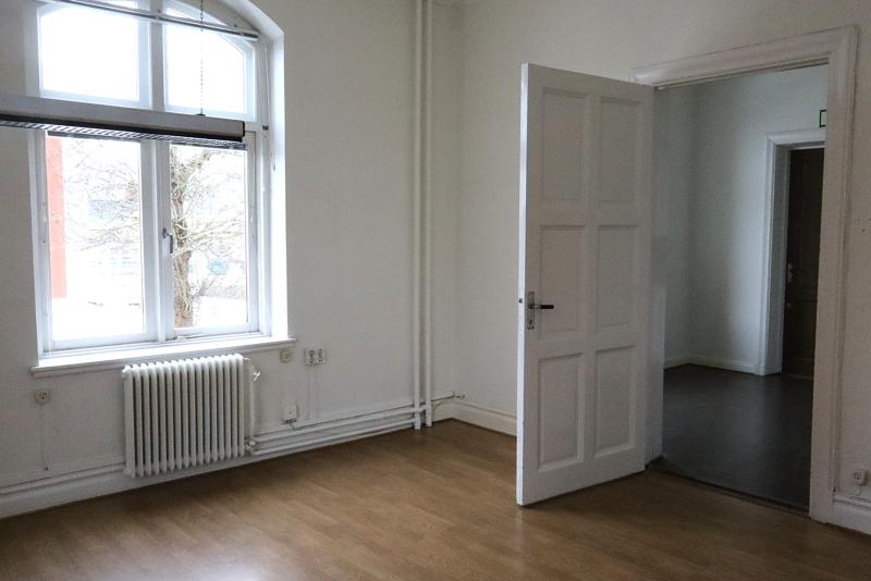 Ett i stort sett tomt rum med ljusa väggar, trägolv, ett stort fönster med ett element under och en öppen vit dörr som leder till ett mörkare angränsande utrymme.