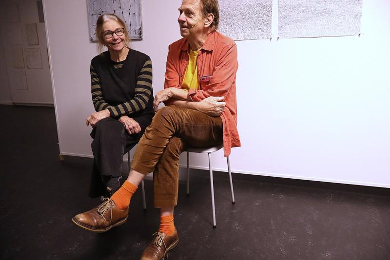 Två personer sitter på stolar i ett starkt upplyst rum med abstrakta konstverk på väggarna; den ena bär en svart klädsel, den andra en orange skjorta, bruna byxor och orange strumpor.