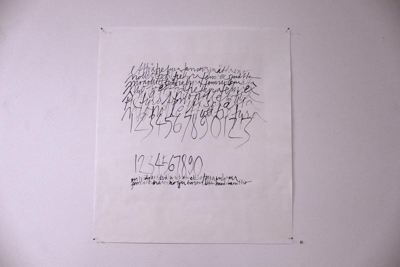 Ett pappersark är fäst på en vägg och visar handskrivna siffror och kursiv text i svart bläck, med viss skrift som verkar klottrad eller överlappande.