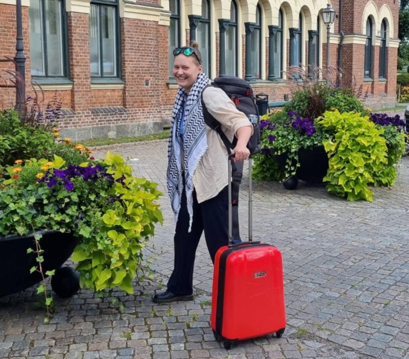 Person som står på en kullerstensgata med en röd resväska och ryggsäck, bredvid stora planteringar med gröna och lila växter, framför en tegelbyggnad med välvda fönster.